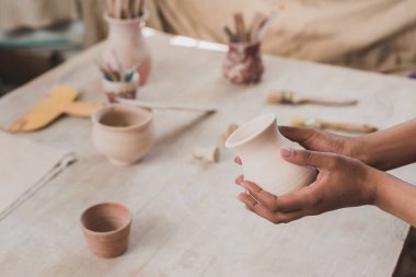 partial view of young african american woman holding clay pot in hands near table with equipment in pottery