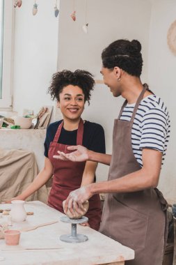 smiling young african american man sculpting clay pot with hand gesturing near woman in pottery