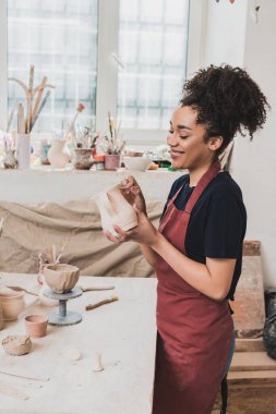 smiling young african american woman sculpting clay pot with stick in pottery