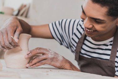 positive young african american man sculpting clay pot with stick in pottery