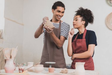 smiling young african american man making clay pot near woman with hand near face in pottery