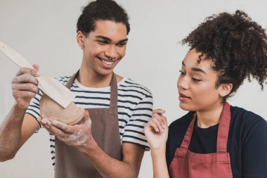 smiling young african american man making clay pot with spatula near woman with hand near face in pottery