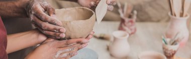 partial view of young african american couple making clay pot with spatula and hands on table with equipment in pottery, banner