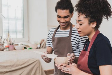 smiling young african american man standing with clay pot and spatula near focused woman in pottery