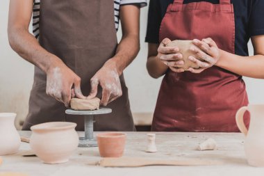partial view of young african american couple in aprons making clay pots on table with equipment in pottery
