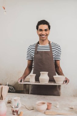 smiling young african american man standing with clay pots on tray in pottery