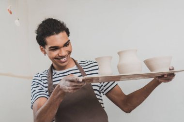 smiling young african american man looking at clay pots on tray in pottery