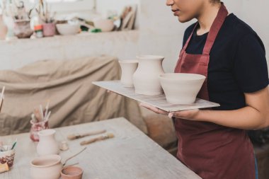 partial view of young african american woman holding clay pots on tray in pottery