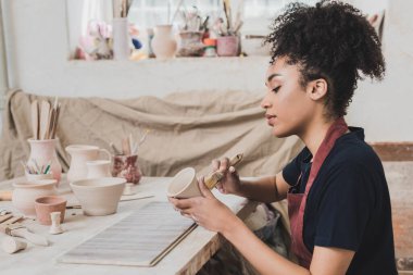 serious young african american woman glazing clay pot with brush near table with equipment in pottery