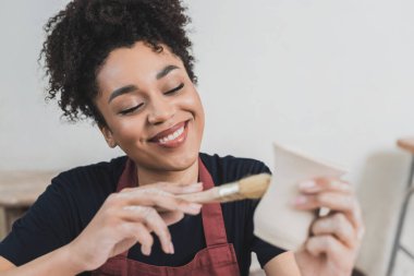 smiling young african american woman glazing clay pot with brush in pottery