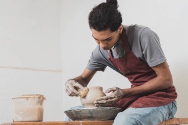 serious young african american man in casual clothes and apron modeling wet clay pot on wheel in pottery