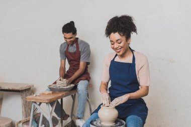 smiling young african american couple in aprons working with wet clay pots in pottery