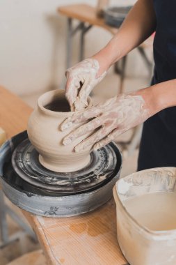 partial view of young african american woman shaping wet clay pot on wheel with hands in pottery