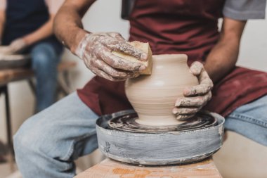 partial view of young african american man holding sponge and making wet clay pot on wheel in pottery