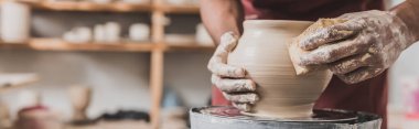 close up view of young african american man holding sponge and making wet clay pot on wheel in pottery, banner