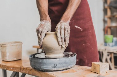 partial view of young african american man shaping wet clay pot on wheel with hands in pottery