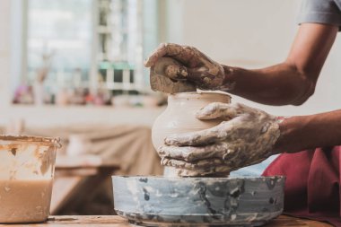 partial view of young african american man in apron sculpting pot on wheel with scraper in hand in pottery