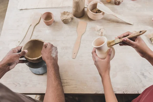 partial view of young african american couple sculpting clay pots with hands and brush in pottery
