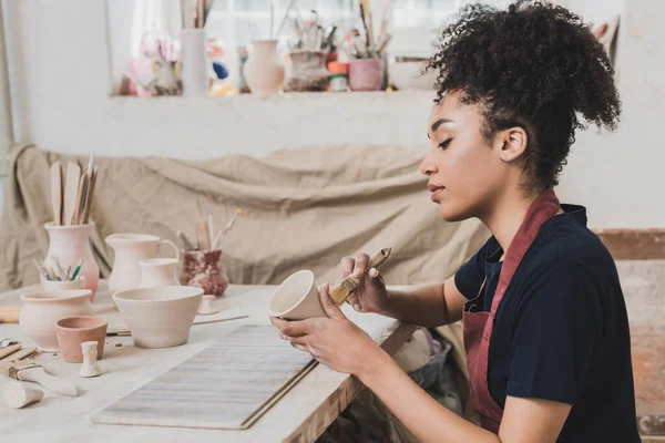严肃的非洲裔美国妇女 用刷子在餐桌边的陶瓷壶上涂釉料 并配有陶器 — 图库照片