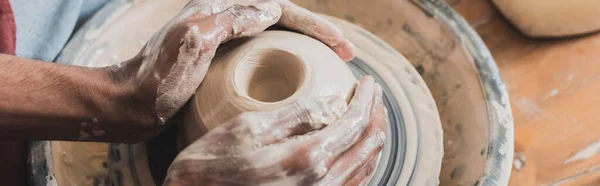 close up view of young african american man modeling wet clay on wheel with hands in pottery, banner