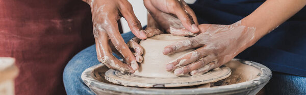 partial view of young african american couple modeling wet clay on wheel with hands in pottery, banner