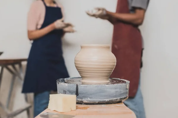 close up view of handmade wet clay pot with young african american couple in aprons on foreground in pottery
