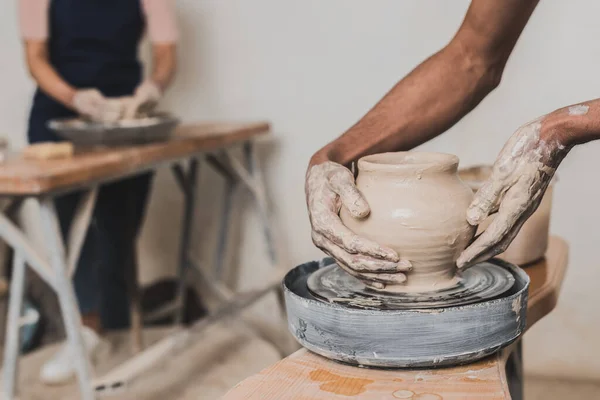 partial view of young african american couple shaping wet clay pots on wheels with hands in pottery