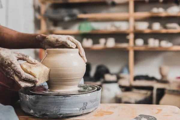 close up view of young african american man holding sponge and making wet clay pot on wheel in pottery