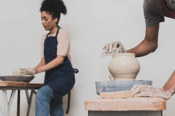 young african american man shaping wet clay pot with scraper on wheel near blurred woman in blue apron in pottery