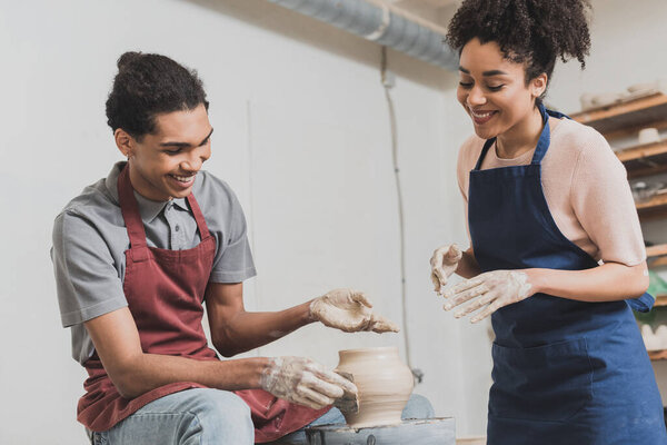 smiling young african american couple shaping wet clay pot on wheel with hands and scraper in pottery