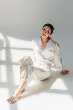 Beyaz elbiseli çıplak ayaklı Asyalı kadın gri arka planda güneş ışığında oturuyor. 