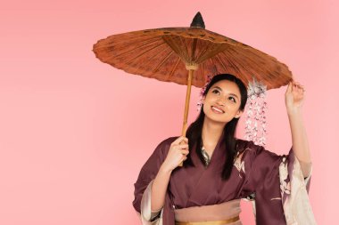 Happy japanese woman in traditional clothes standing under umbrella isolated on pink  clipart
