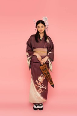 Japanese woman with traditional hairdo and kimono holding umbrella on pink background clipart