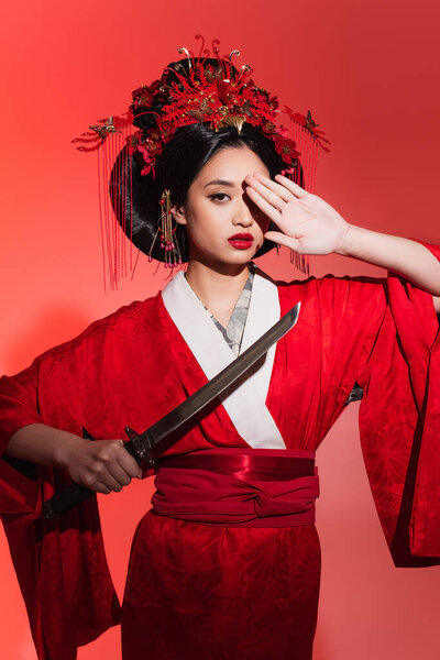 Asian woman in oriental clothes holding sword and covering eye on red background 