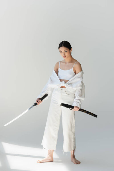 Barefoot japanese woman in white clothes holding sword on grey background 