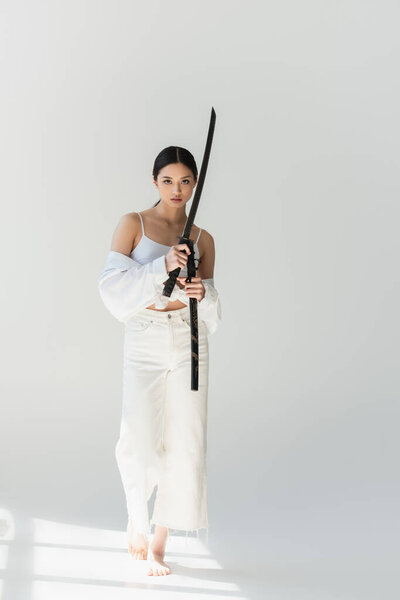 Barefoot asian woman with sword walking on grey background with sunlight  