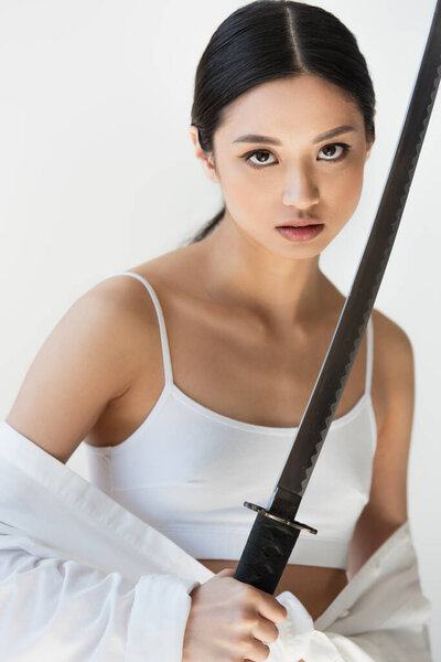 Young asian woman in white top and shirt holding sword isolated on grey