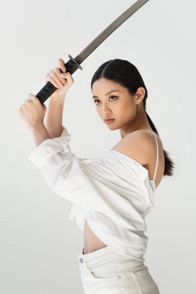 Asian woman in shirt holding sword and looking away isolated on grey