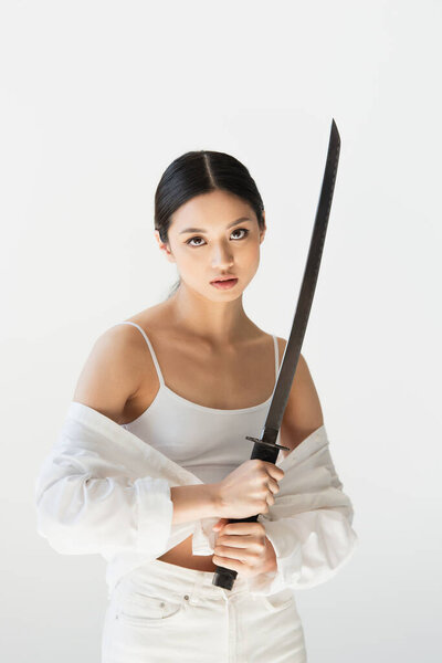 Asian woman in white clothes looking at camera while holding sword isolated on grey