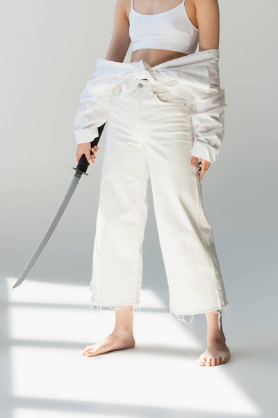 Cropped view of woman in white clothes holding sword on grey background 