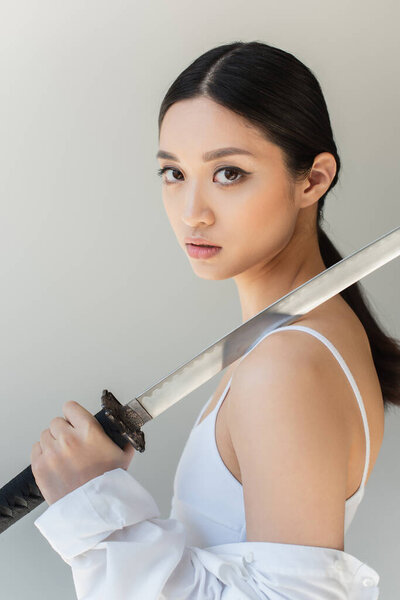 Asian woman in white clothes holding sword on shoulder isolated on grey