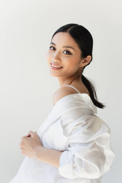 Pretty asian woman adjusting white shirt and smiling at camera isolated on grey