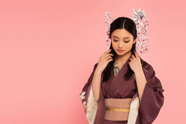 Young asian woman in kimono adjusting brunette hair isolated on pink 