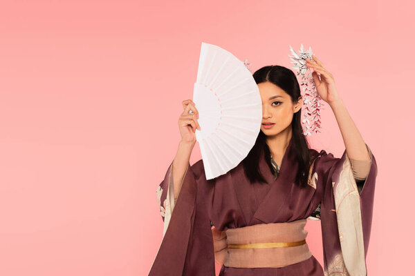 Asian woman with traditional kanzashi in hair covering face with fan isolated on pink 