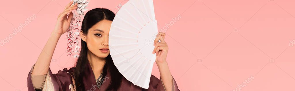 Asian woman with traditional hairdo holding fan near face isolated on pink, banner 