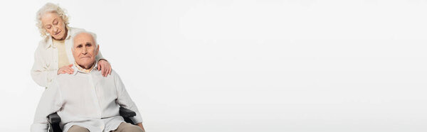 elderly woman holding hands on shoulders of husband in wheelchair isolated on white, banner