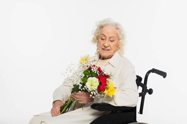 smiling elderly woman in wheelchair holding bouquet of flowers isolated on white