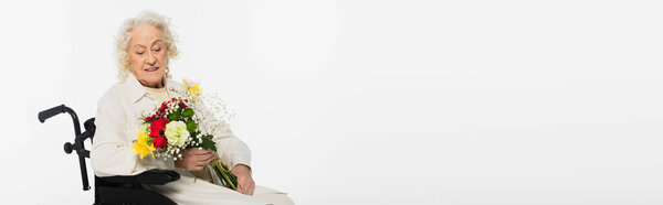 smiling elderly woman in wheelchair holding bouquet of flowers isolated on white, banner