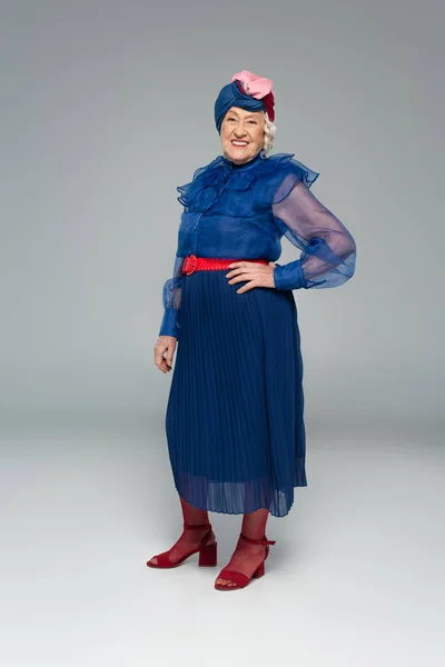 Улыбающаяся Пожилая Женщина Синем Платье Тюрбан Позирующая Рукой Бедре Сером — стоковое фото