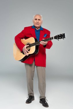 Kırmızı ceketli ve kahverengi pantolonlu yaşlı bir adam gri üzerinde akustik gitar tutuyor.